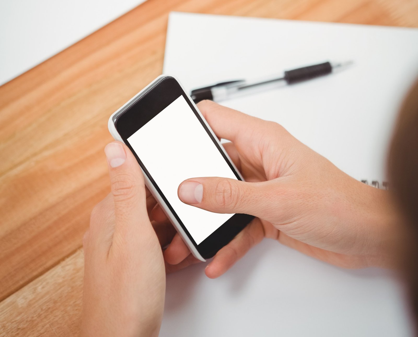 Two hands hold a phone with a blank screen over a wooden table, with a pen and paper beside it.